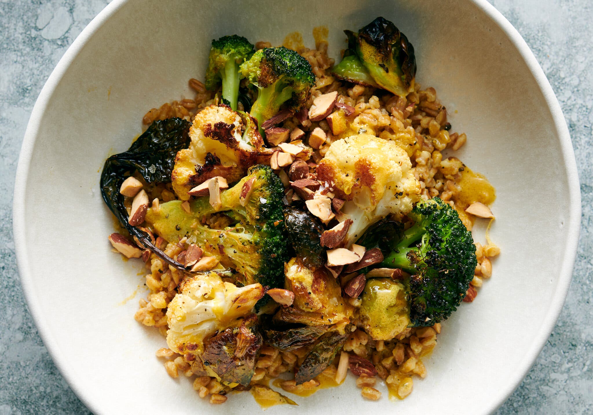 A white ceramic bowl holds roasted broccoli and cauliflower, grains and chopped almonds.