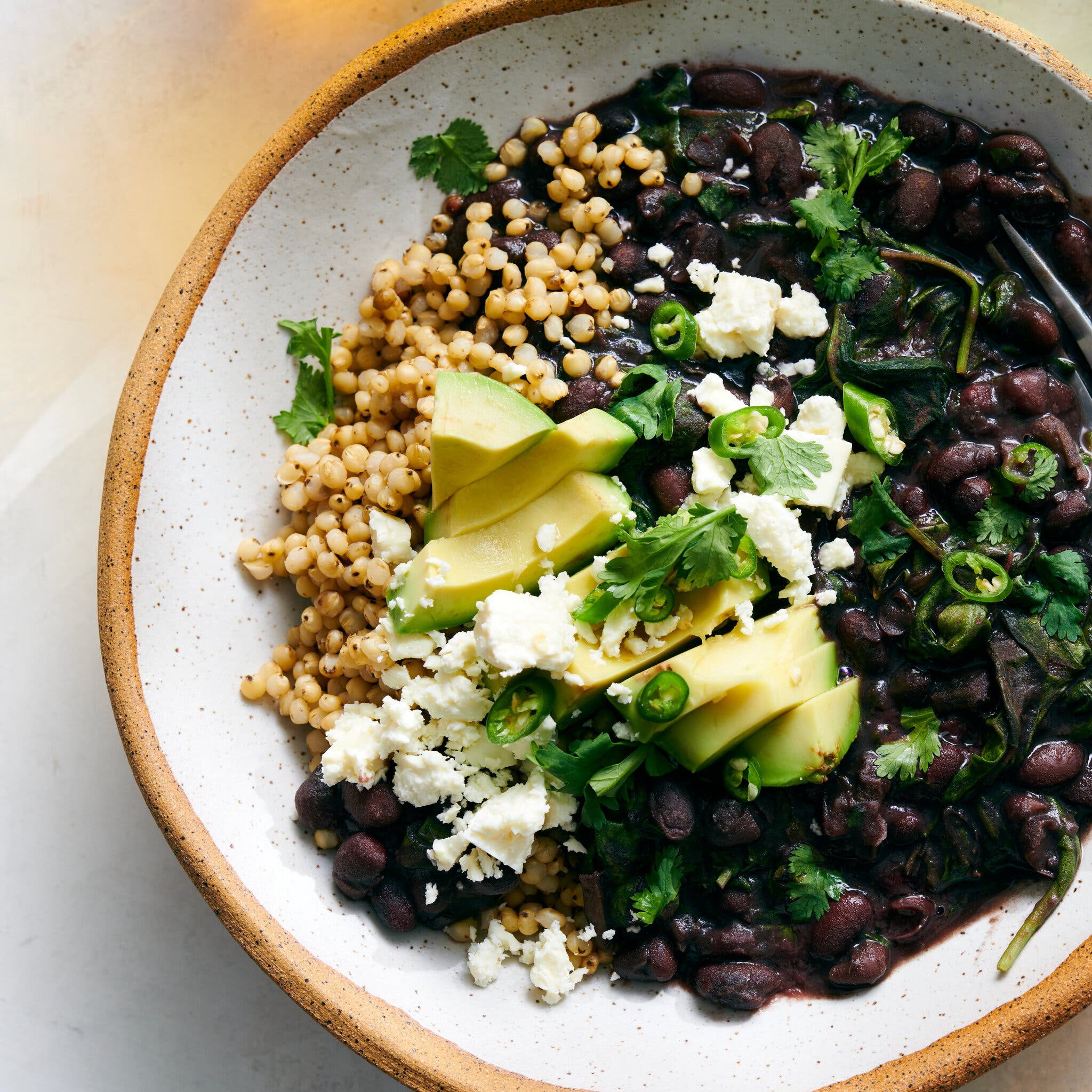 A ceramic bowl holds sorghum, black beans, avocado, cilantro and feta.