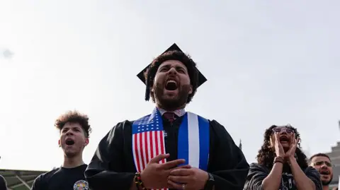 Bloomberg via Getty Images A Harvard University student chants with fellow demonstrators during a protest in support of international students at Harvard University. He is wearing a graduation cap and gown with a sash which has an American flag on it.