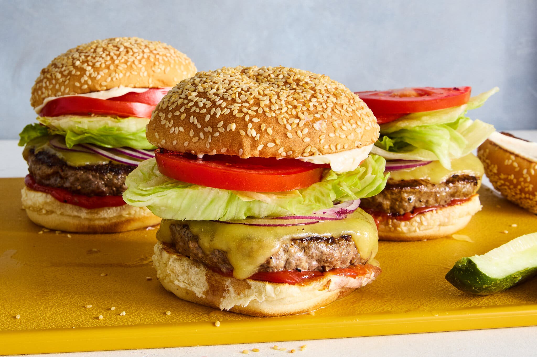 Three burgers on a cutting board topped with cheese, onion, lettuce and tomato.