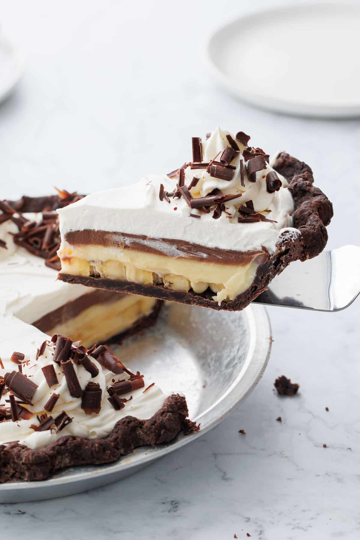 Lifting a slice of Chocolate Banana Cream Pie out of a pie plate on a white marble background.