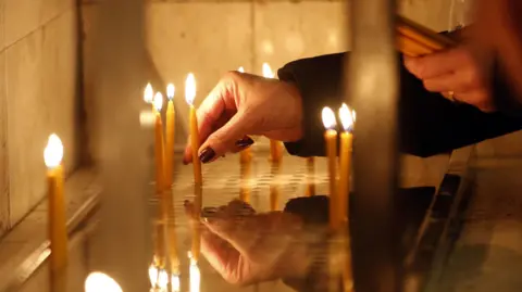 EPA A woman's hand is holding a lit candle and placing it in a row of candles, in what appears to be a church. 